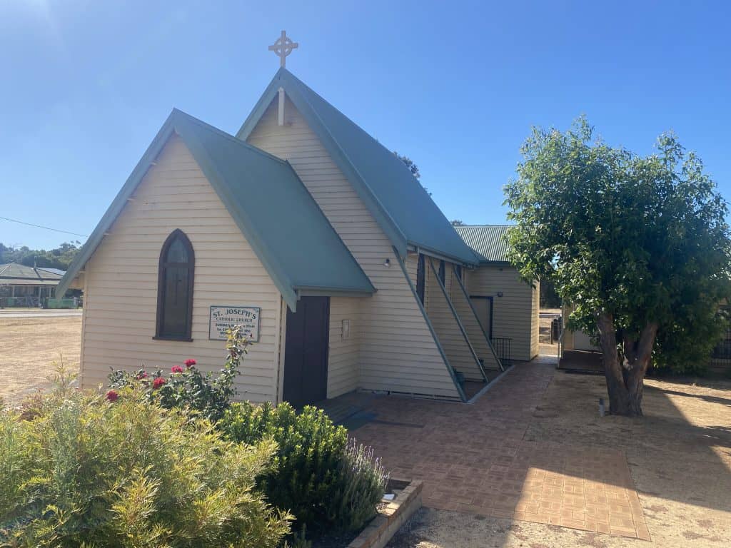 Historic church building with a steep roof and cross, surrounded by garden and trees.