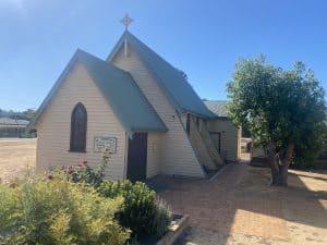 Historic church building with a steep roof and cross, surrounded by garden and trees.