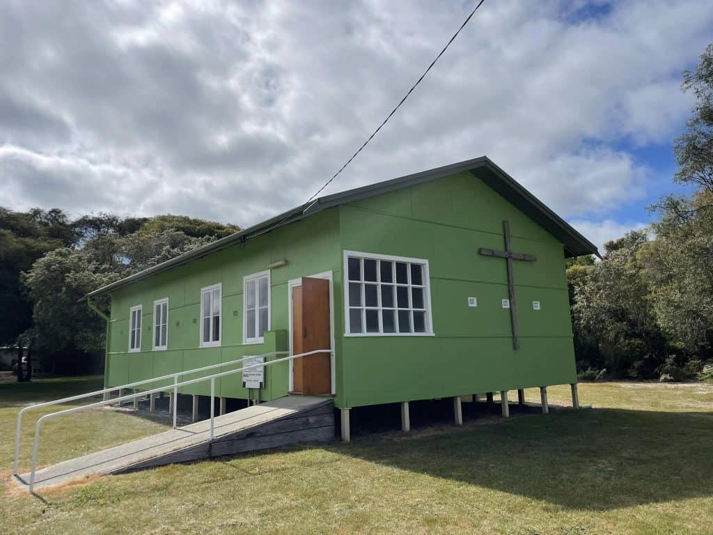 Green church building with cross and ramp in a grassy area under cloudy sky.