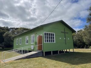 Green church building with cross and ramp in a grassy area under cloudy sky.